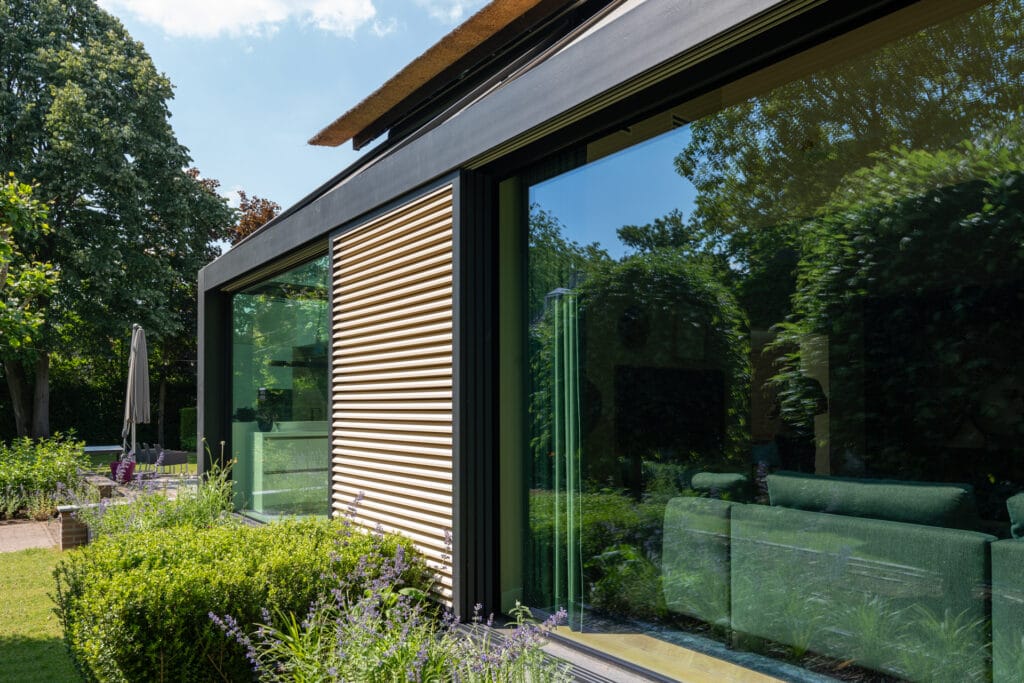 Geautomatiseerde zonwering: antraciete shutters, beige lamellen; Uitbouw met staal en glas, HR++ glaspanelen.