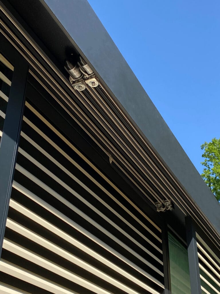 Geautomatiseerde zonwering: antraciete shutters, beige lamellen, mechanisme achter stalen H-ligger.