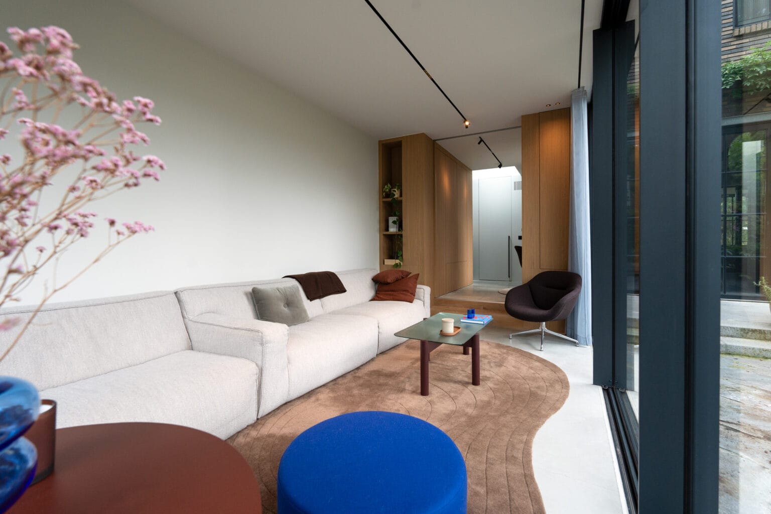 Interieur tuinkamer: glazen hefschuifpui, lang bankstel, afsluitbare houten scheidingswand met ensuite deuren, organisch aardekleurig tapijt, geïntegreerde plafondverlichting.